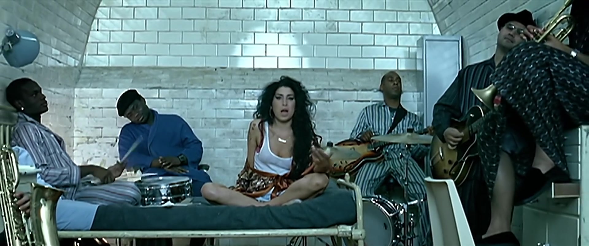 Amy Winehouse in een wit hemdje met haar kenmerkende haar en eyeliner, met haar band