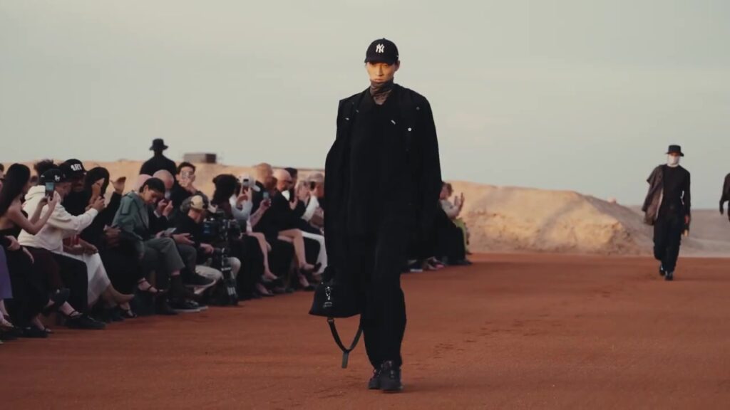 Model met een pet van de Yankees en een lange zwarte jas op een catwalk in de woestijn